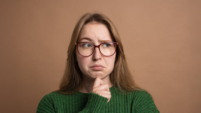 Pensive woman in green sweater looks uncertain or confused, touching chin and raising eyebrows.