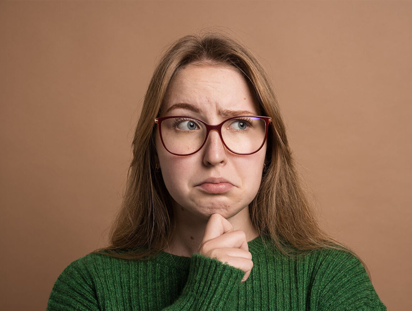 Pensive woman in green sweater looks uncertain or confused, touching chin and raising eyebrows.