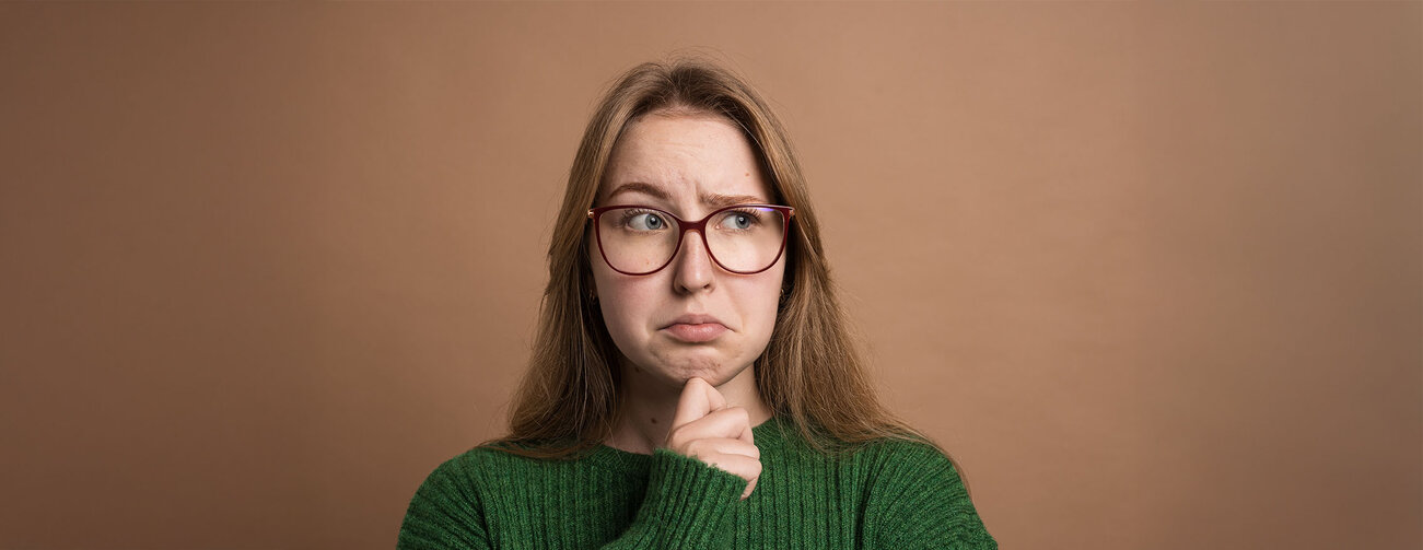 Pensive woman in green sweater looks uncertain or confused, touching chin and raising eyebrows. 