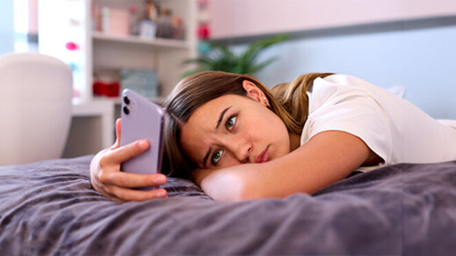 Depressed Teenage Girl Lying On Bed At Home Looking At Mobile Phone